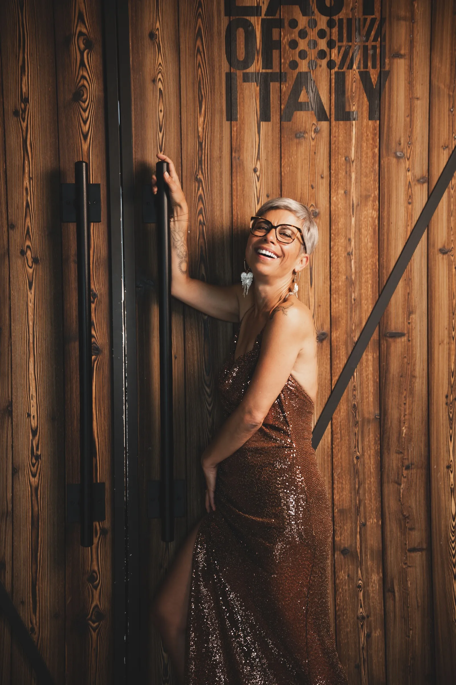 Doritt Adam lacht in einem goldfarbenen Kleid vor einer rustikalen Holzwand.